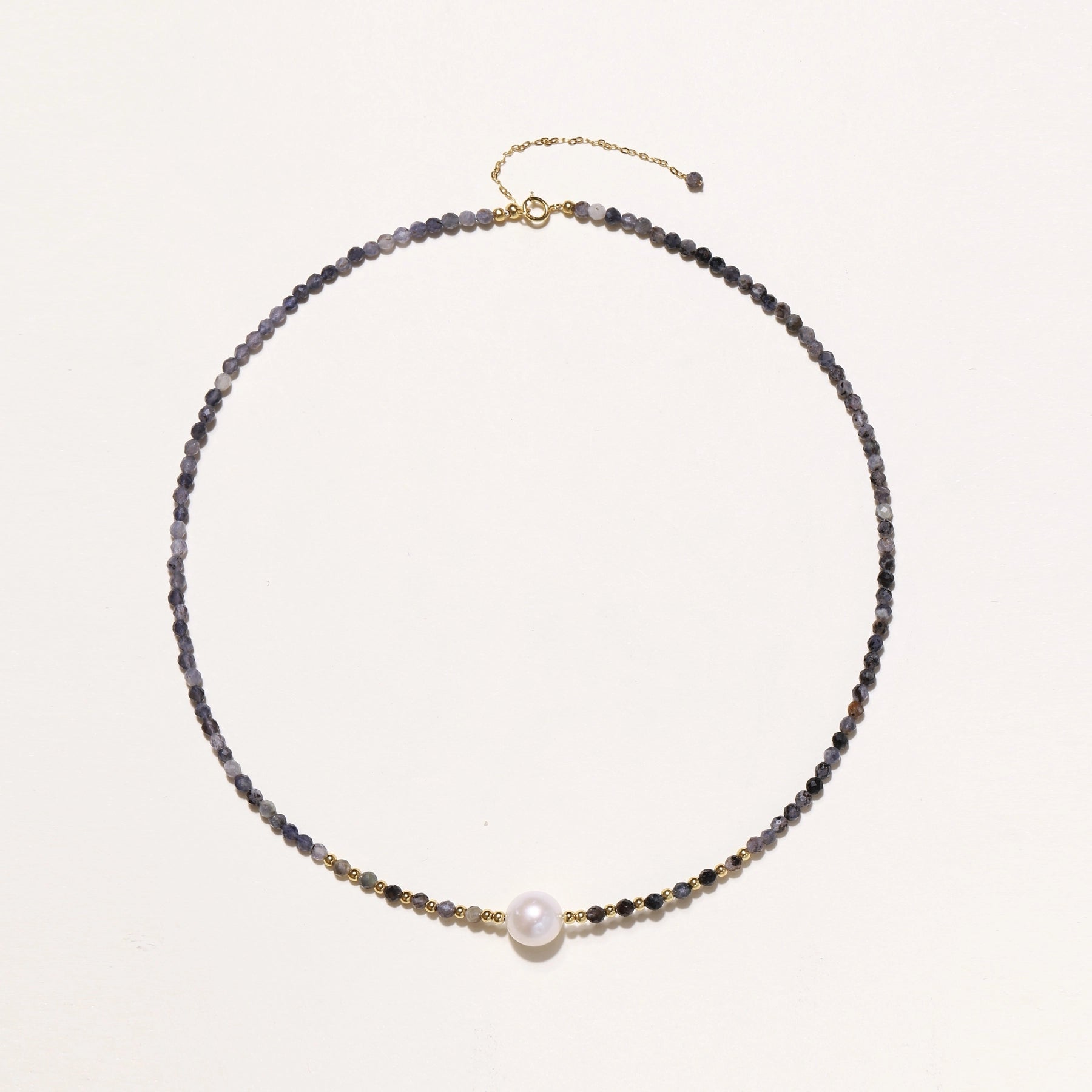 Delicate beaded necklace with violet iolite stones, gold spacers, and central white freshwater pearl on neutral background.