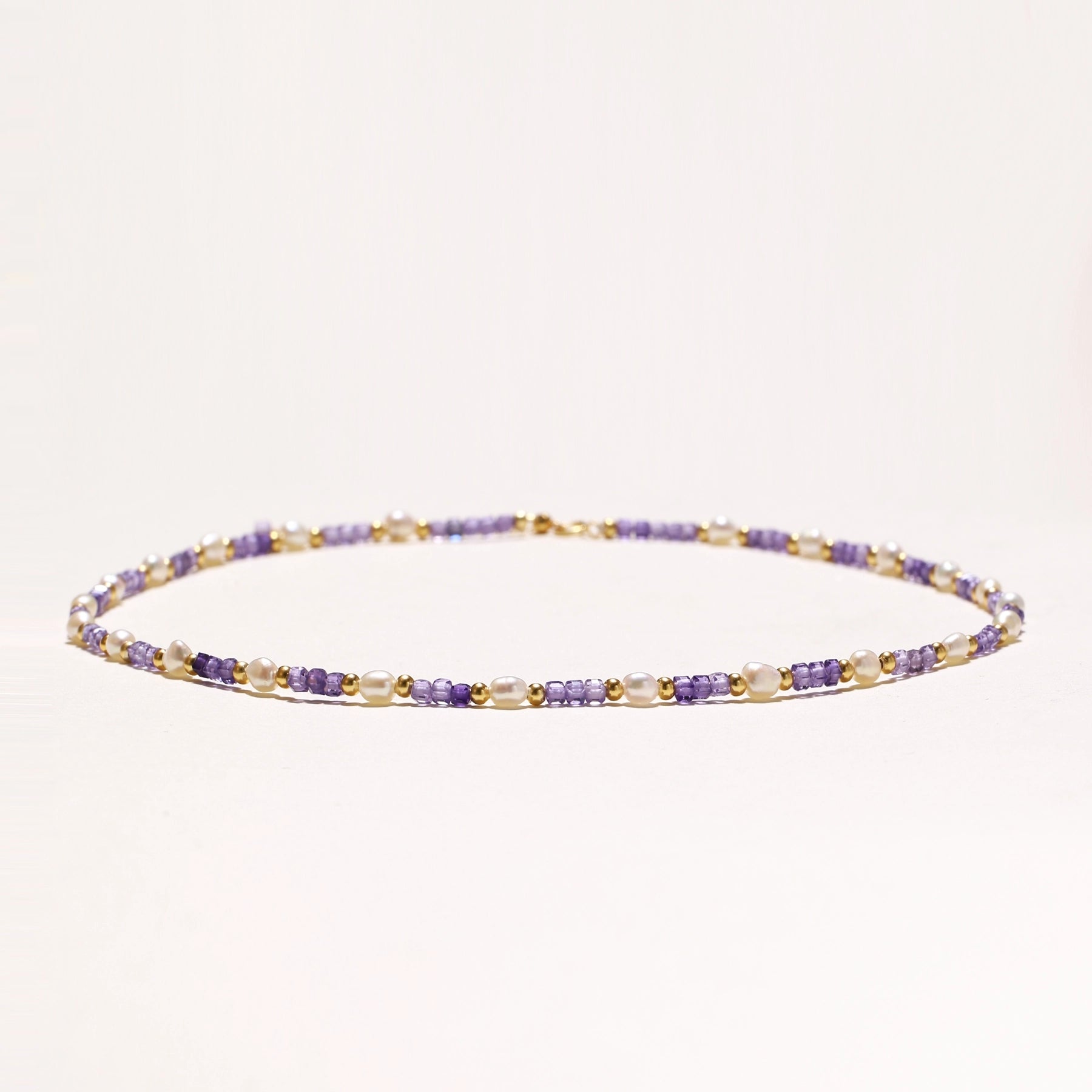 Delicate beaded necklace with alternating purple zirconia stones, white freshwater pearls, and gold-plated spacers on cream background.