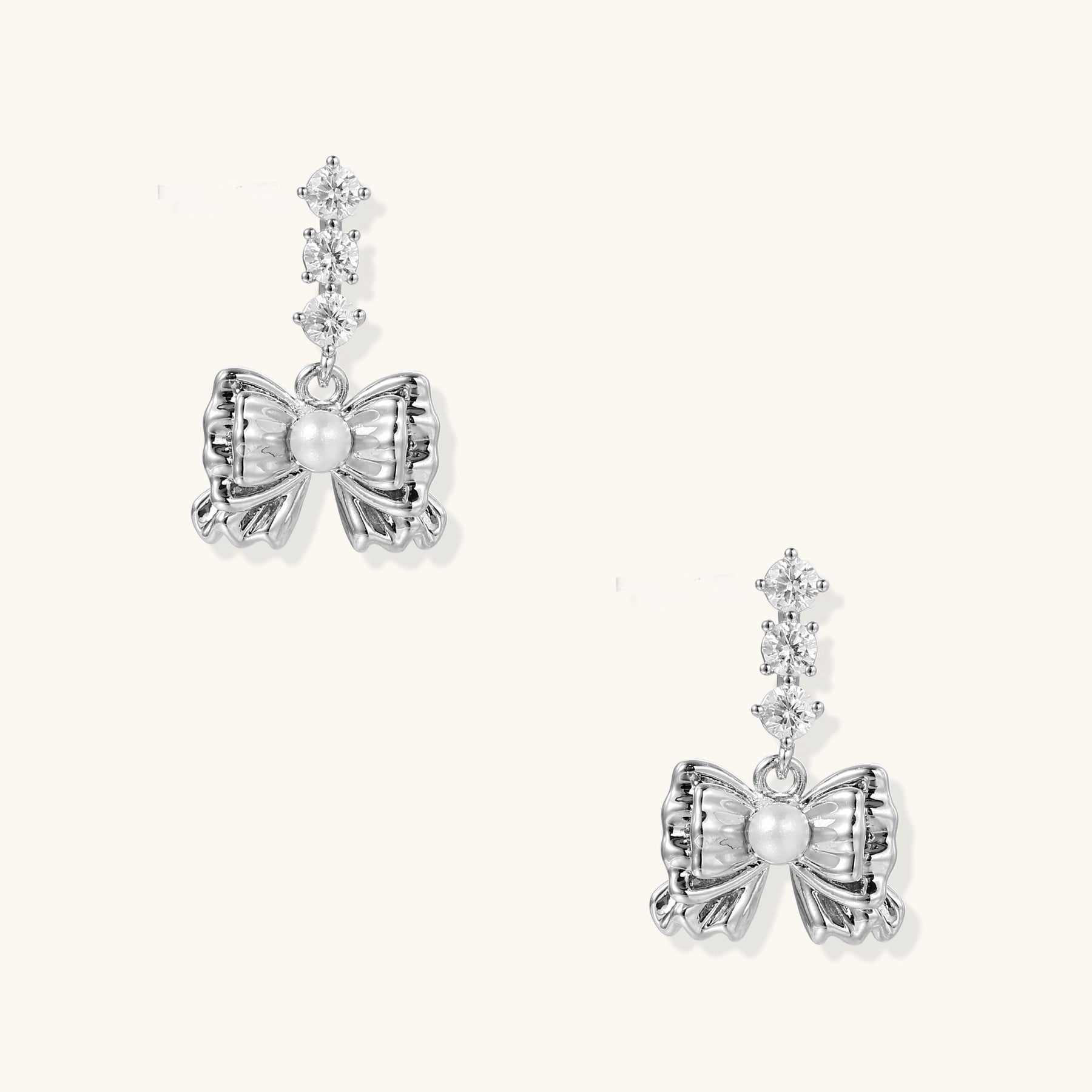 Delicate gold-plated bow drop earrings with three zirconia crystals and center pearl on white background.